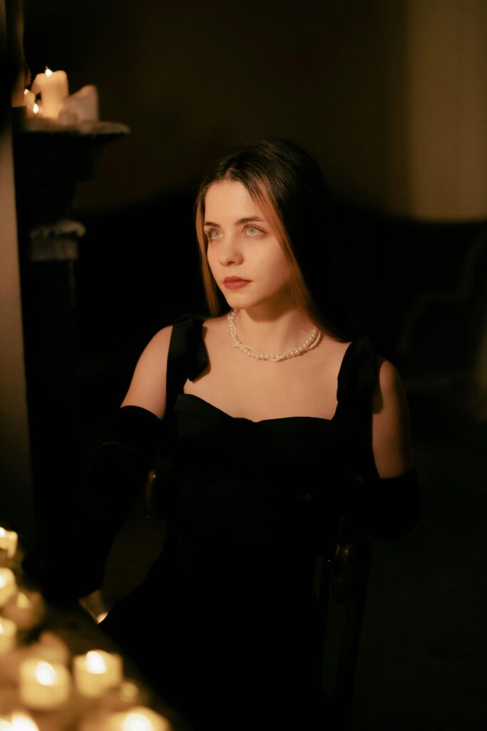 A sophisticated woman in black dress and pearls illuminated by candlelight in an intimate setting.