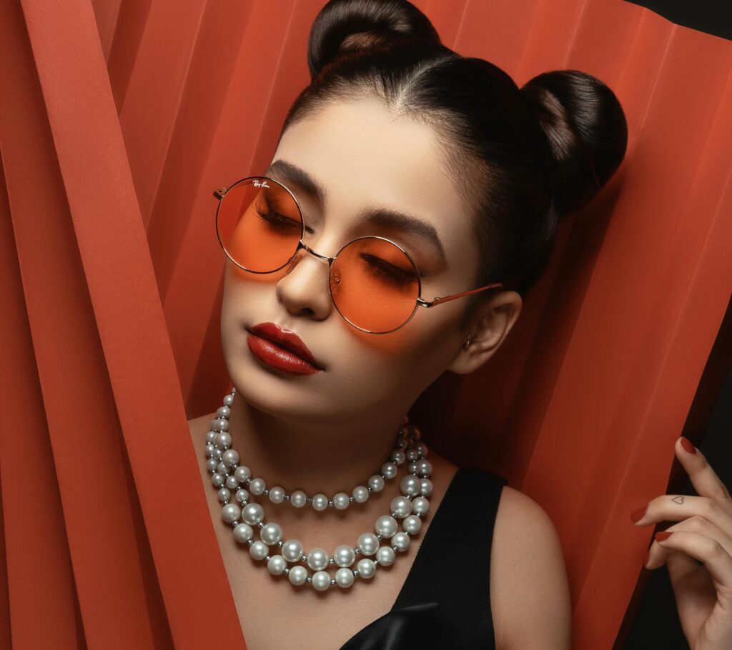 Stylish woman in black dress, pearls, and orange sunglasses posing gracefully.